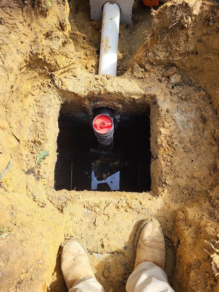 Open utility hole with a white pipe and red-capped pipe, viewed from a dirt trench with boots at bottom.