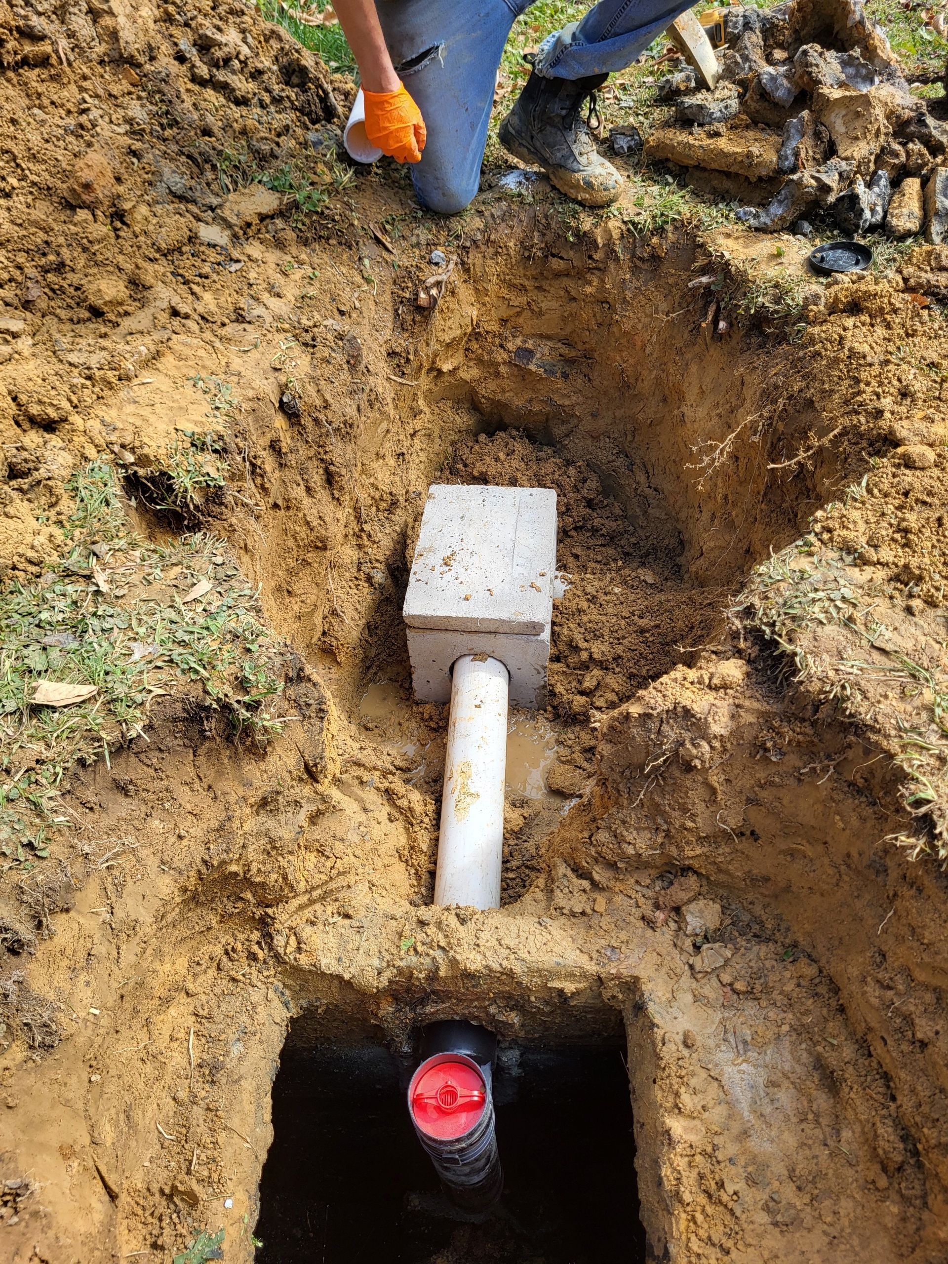 Worker digging a trench with a buried pipe and utility box; red pipe opening visible underground