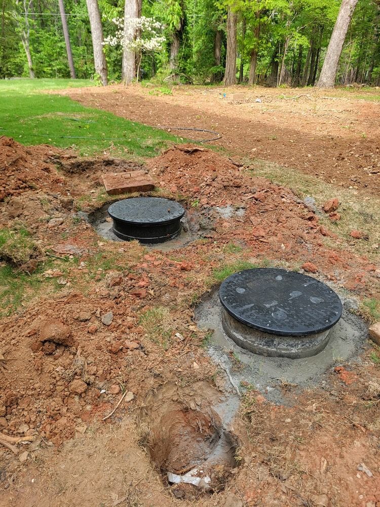 Two black circular utility lids set in fresh dirt beside a wooded path.
