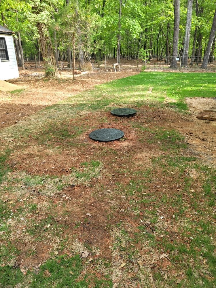 Two green utility lids in a grassy dirt yard near trees and a white building.