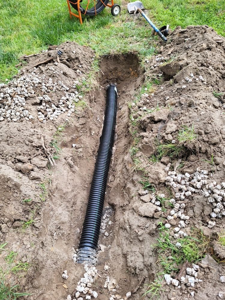 Trench with black drainage pipe laid in soil, surrounded by excavated dirt and grass.