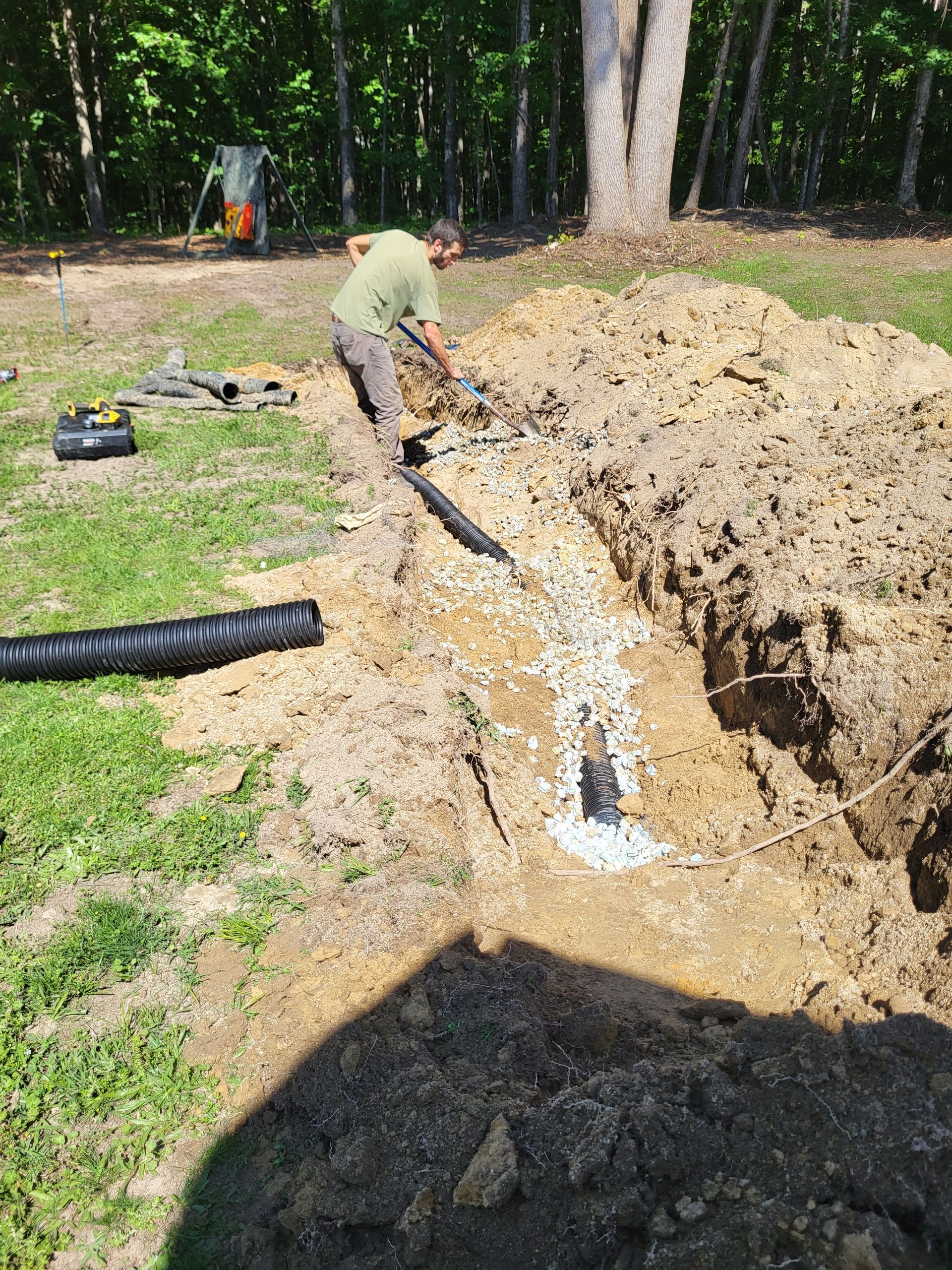 Worker digging a trench beside a wooded yard, with a black drainage pipe in the grass.