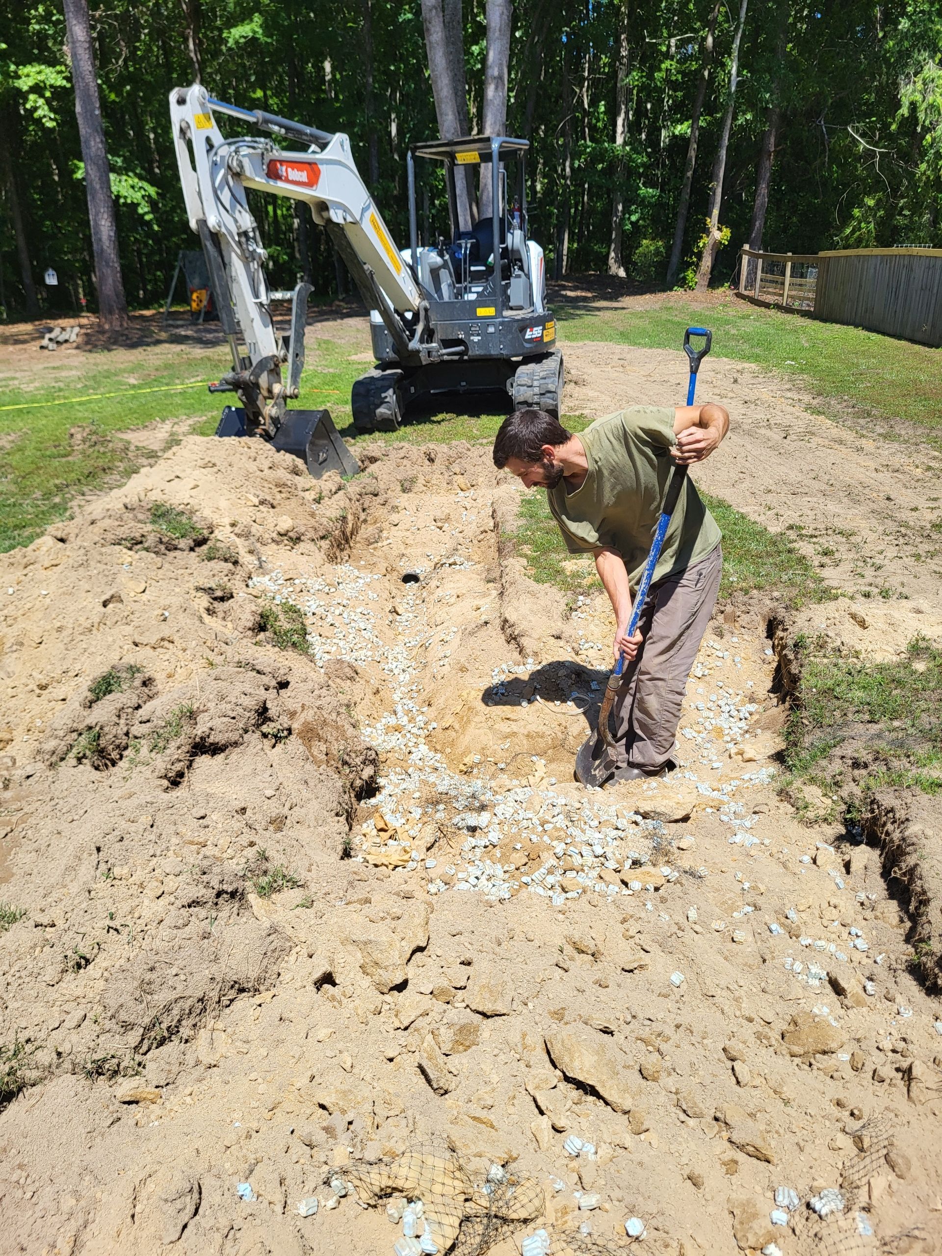 Person digging in a large dirt trench beside a small excavator in a wooded yard
