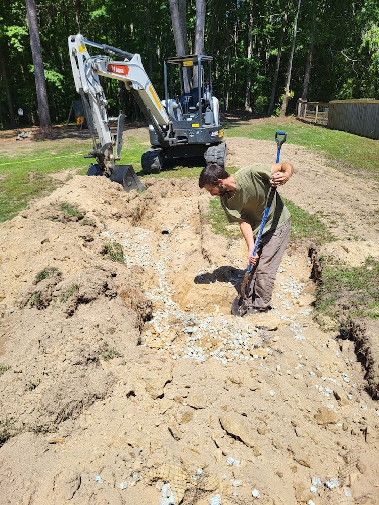 Worker shoveling muddy trench beside an excavator in a wooded yard