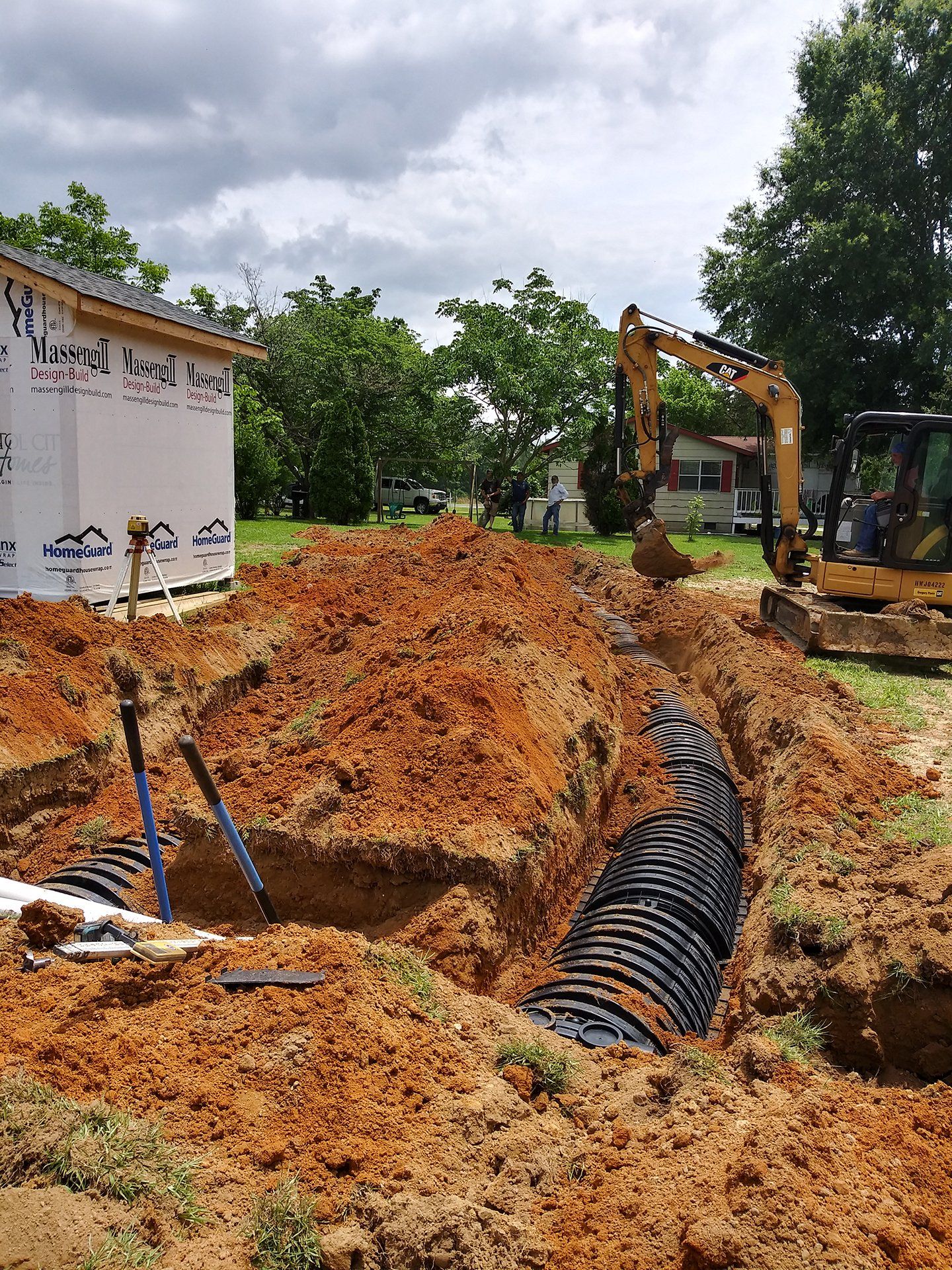 Construction trench with black drainage pipe beside a small building and excavator under cloudy sky