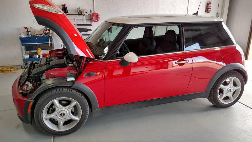 A red Mini Cooper with a white roof parked inside a garage with its hood open, revealing the engine bay.