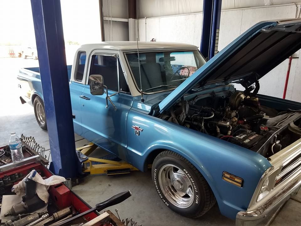 A light blue and white classic pickup truck with its hood open, parked in an auto repair shop on a vehicle lift.