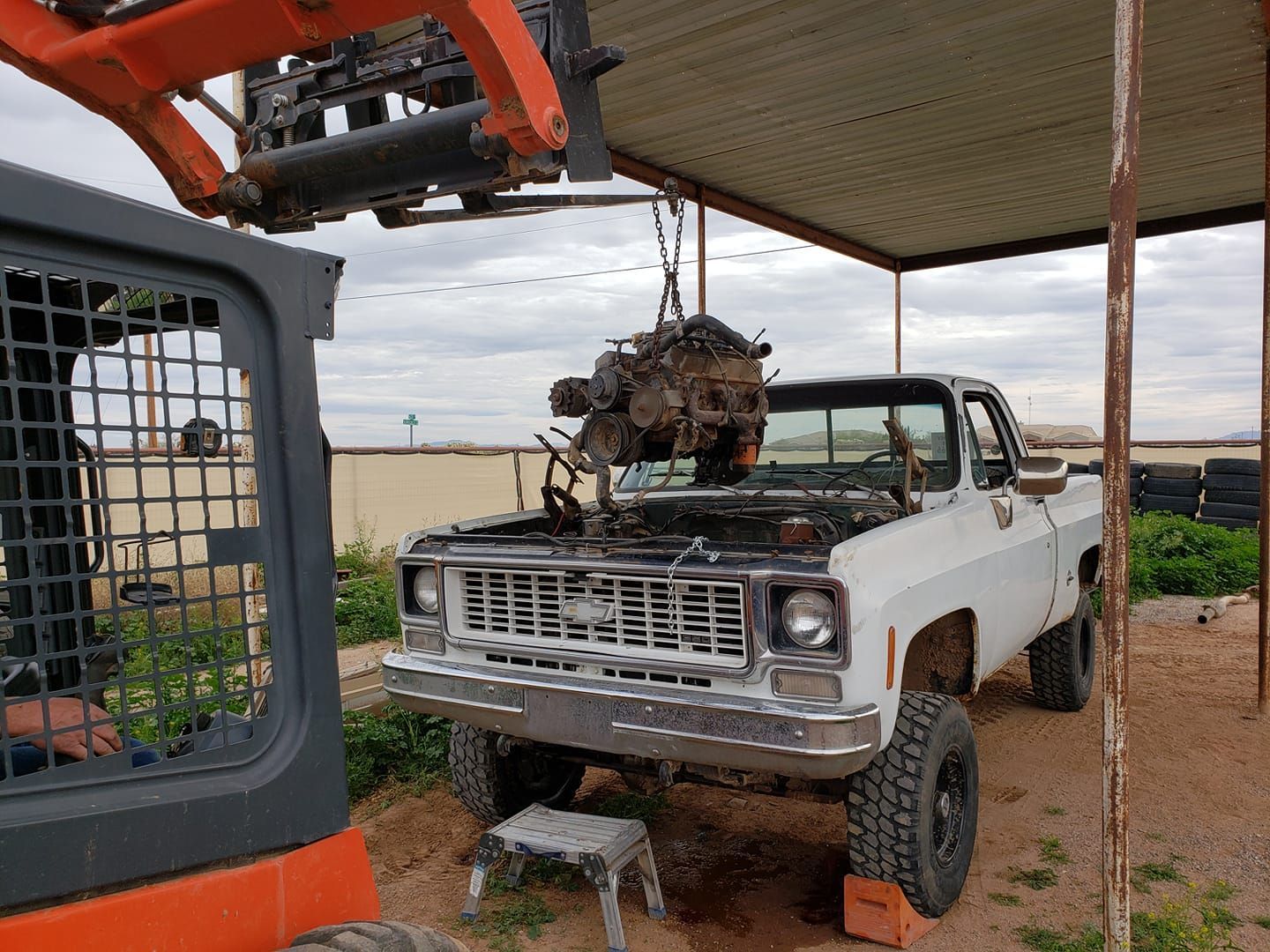 A skid steer loader lifts an engine out of the open hood of a white pickup truck parked under a covered outdoor structure.