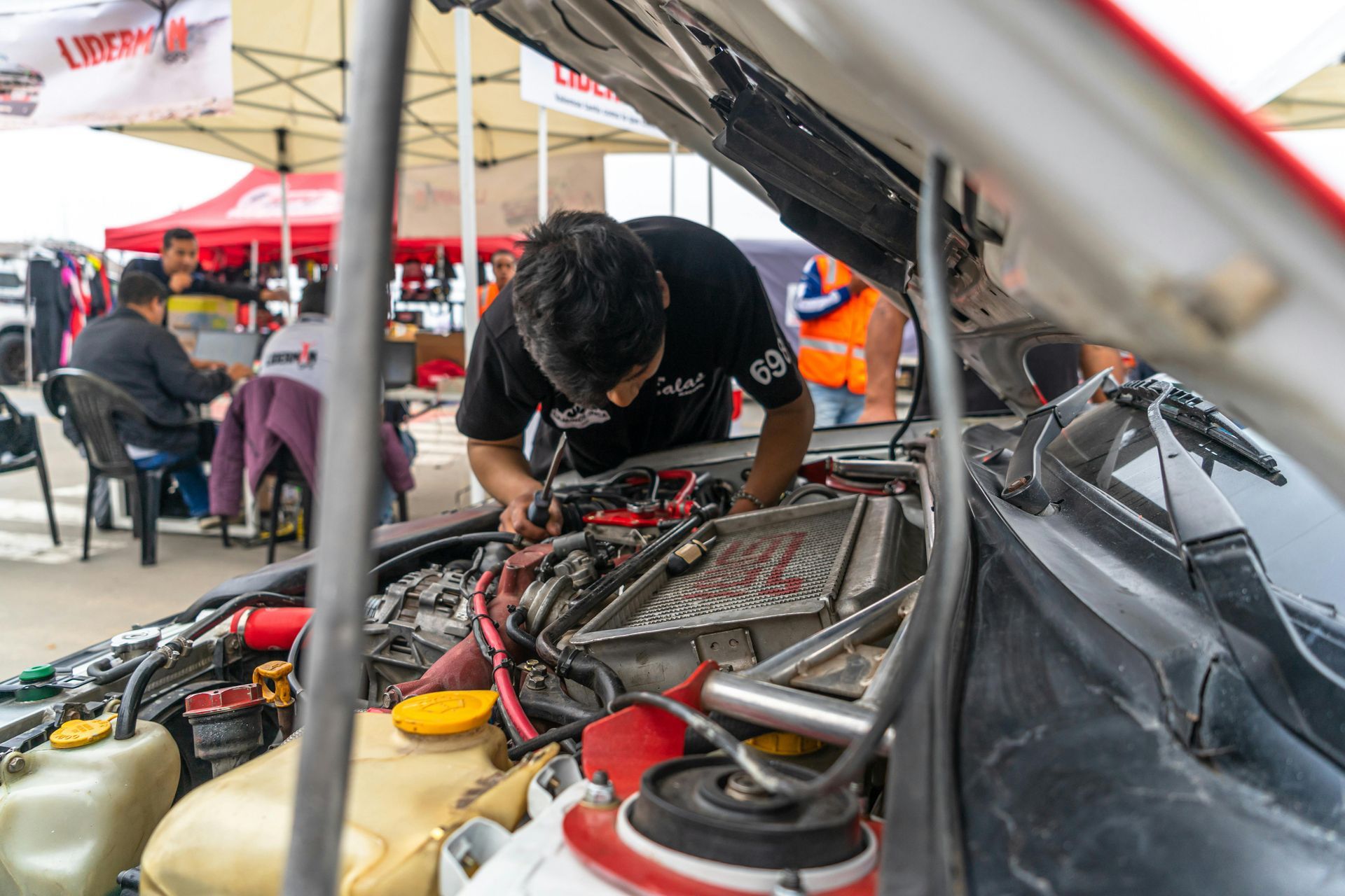 A person in a black shirt works on the engine of a vehicle under an outdoor canopy at a racing or repair event.