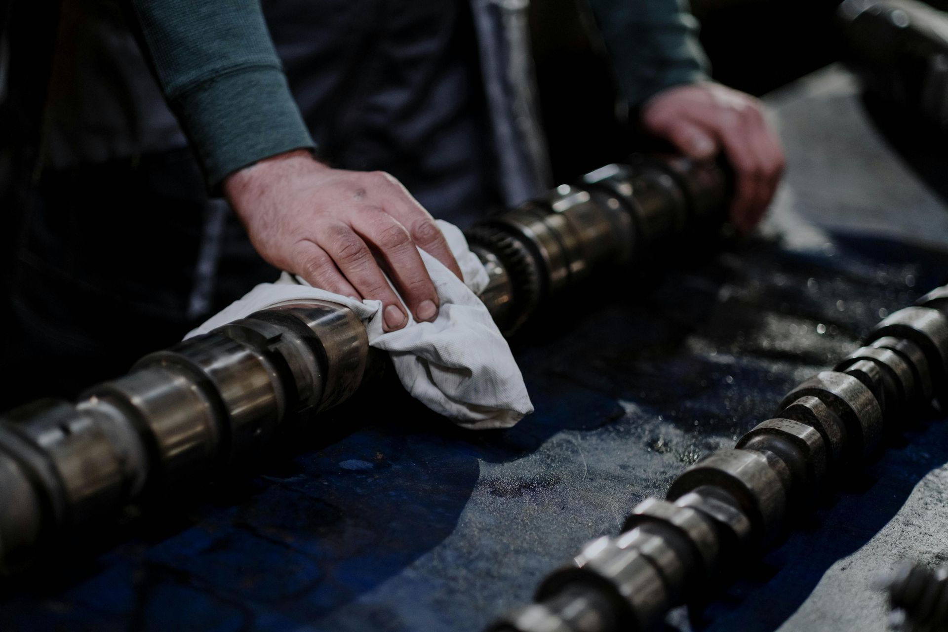 Hands wearing a green sleeve wipe down a metal engine camshaft with a white cloth in a workshop.