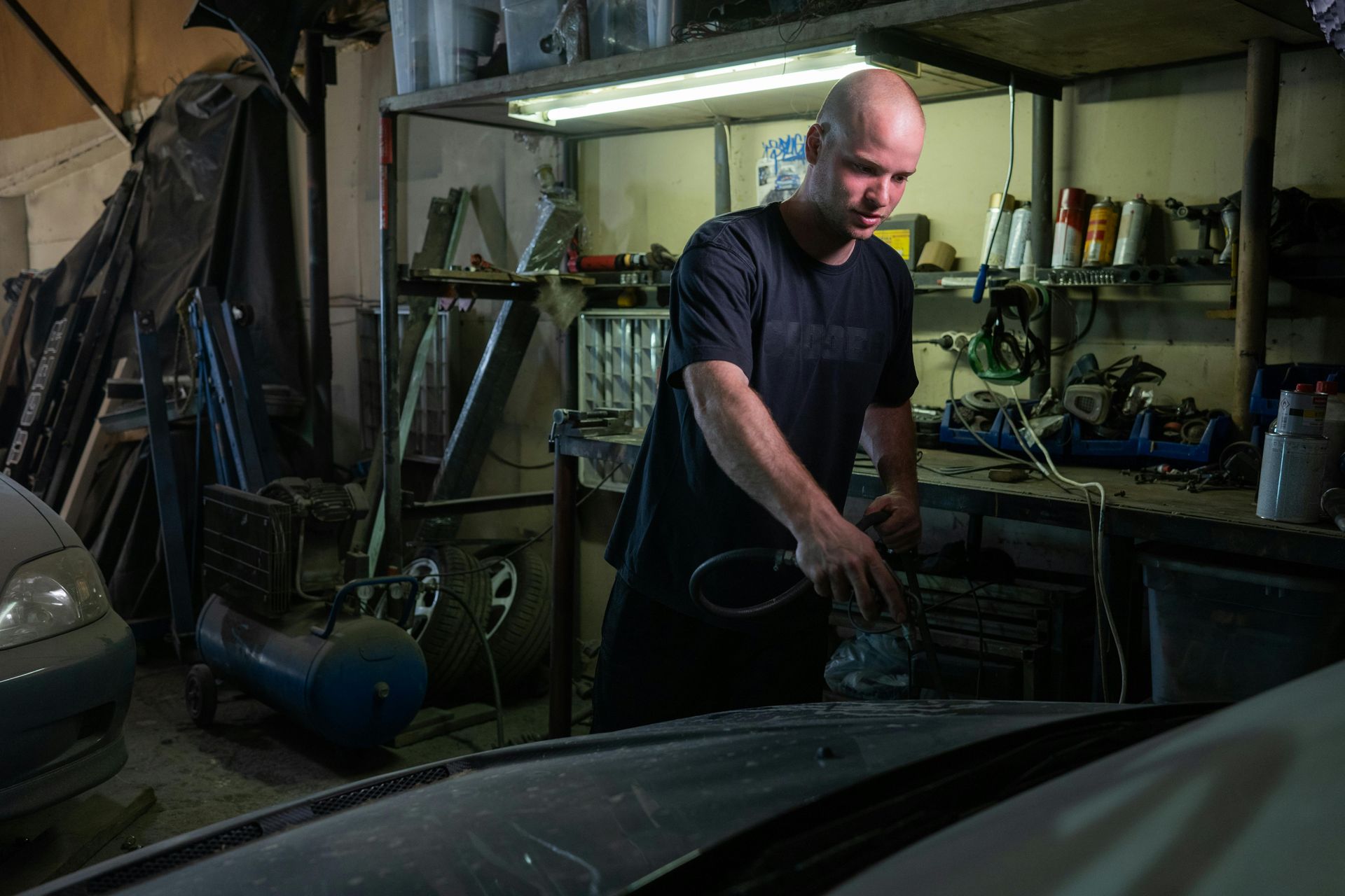 A person in a dark shirt uses a sander on a car hood inside a dimly lit workshop filled with tools and parts.