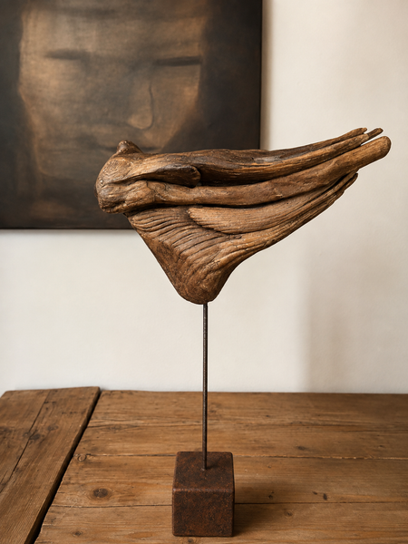 Scultura astratta in legno su un supporto metallico appoggiato su un tavolo di legno, con un'opera d'arte incorniciata di scuro sullo sfondo.