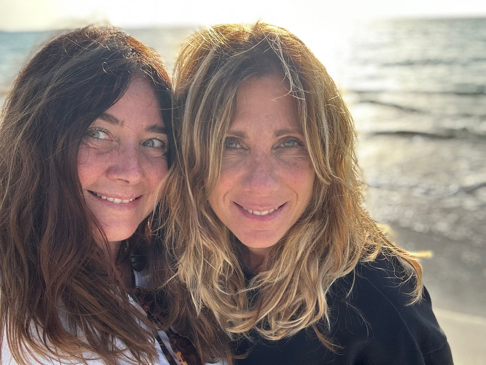Due donne sorridenti con i capelli scompigliati dal vento posano su una spiaggia assolata.