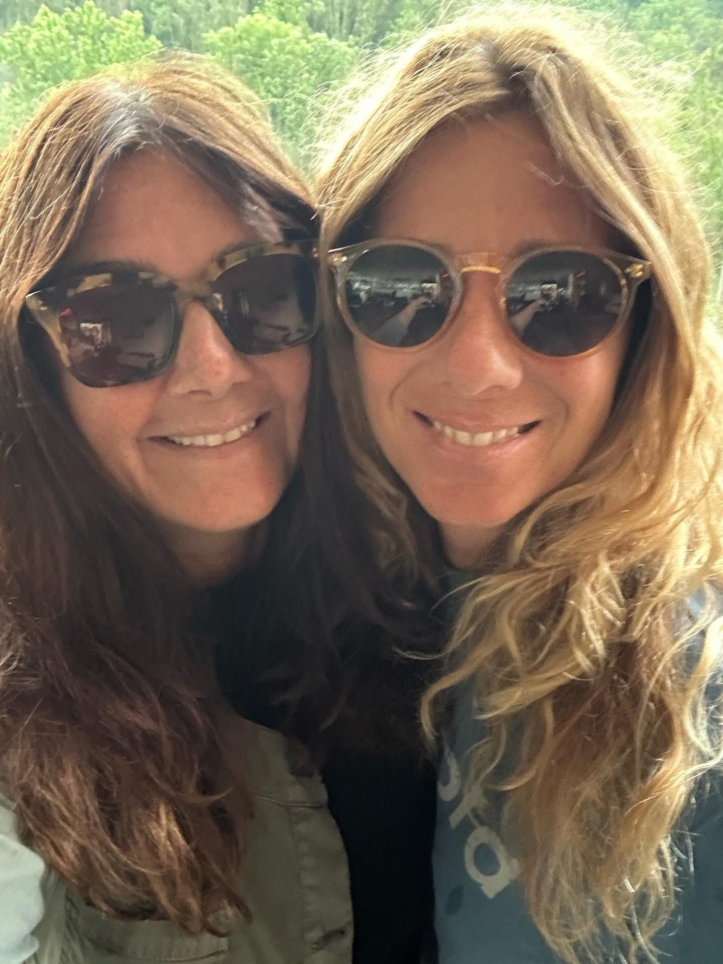 Due donne sorridenti con occhiali da sole posano all'aperto sotto la luce del sole.