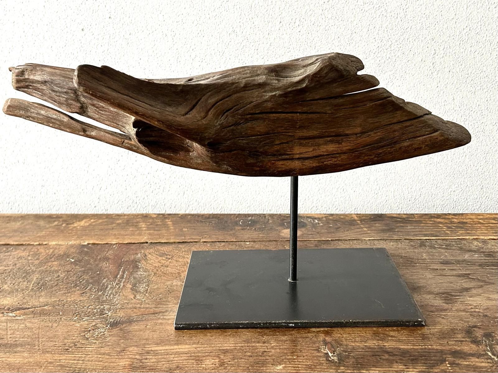 Scultura astratta in legno su supporto metallico con base nera, su sfondo di una parete bianca.