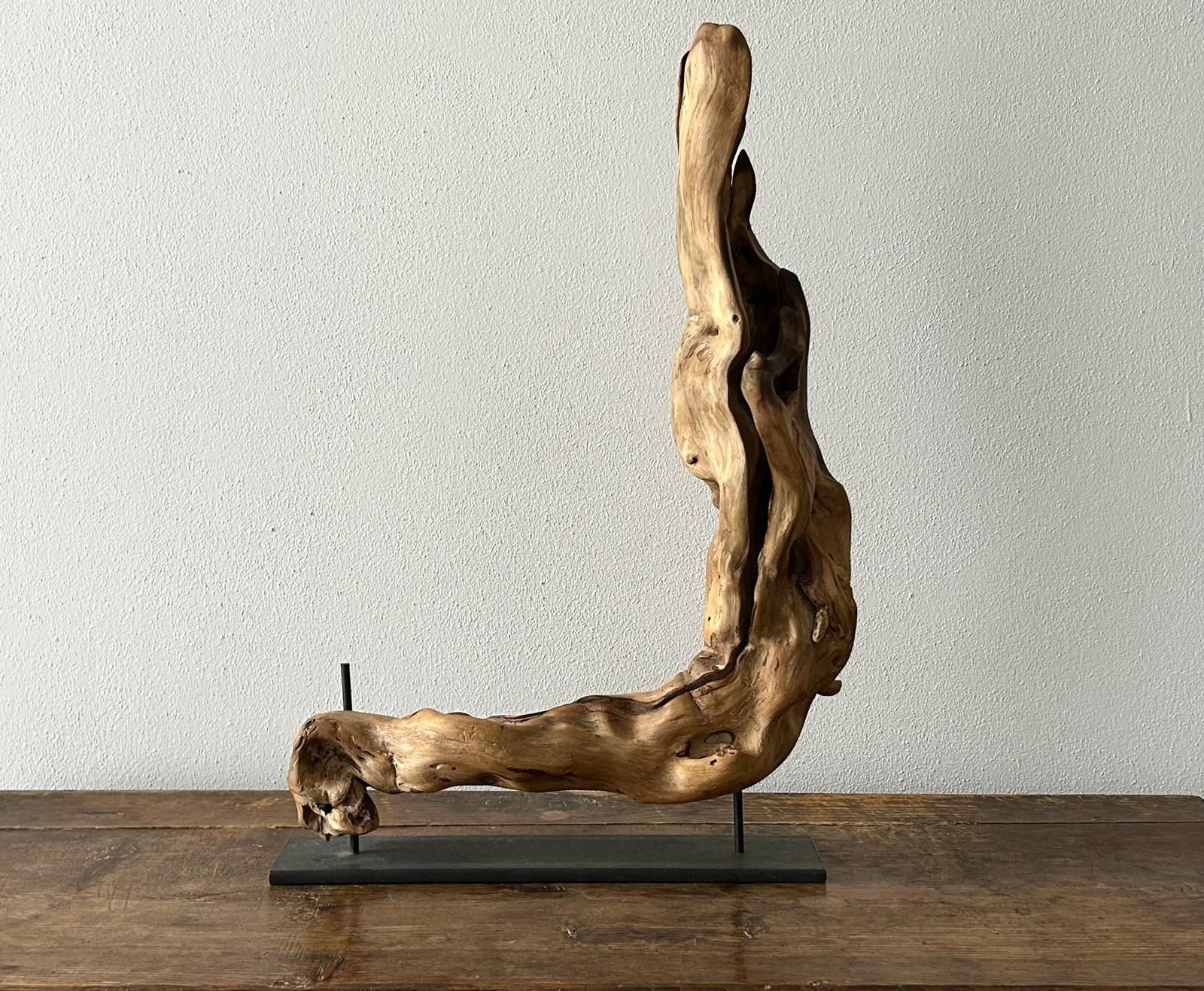 Scultura astratta in legno di recupero, poggiata su un supporto in metallo nero, contro una parete spoglia.