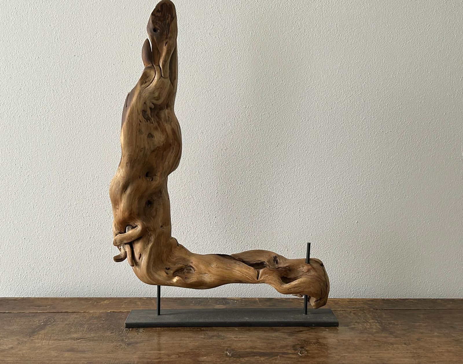 Scultura astratta in legno raffigurante una figura umana sdraiata su una base nera, sullo sfondo di una parete grigia.