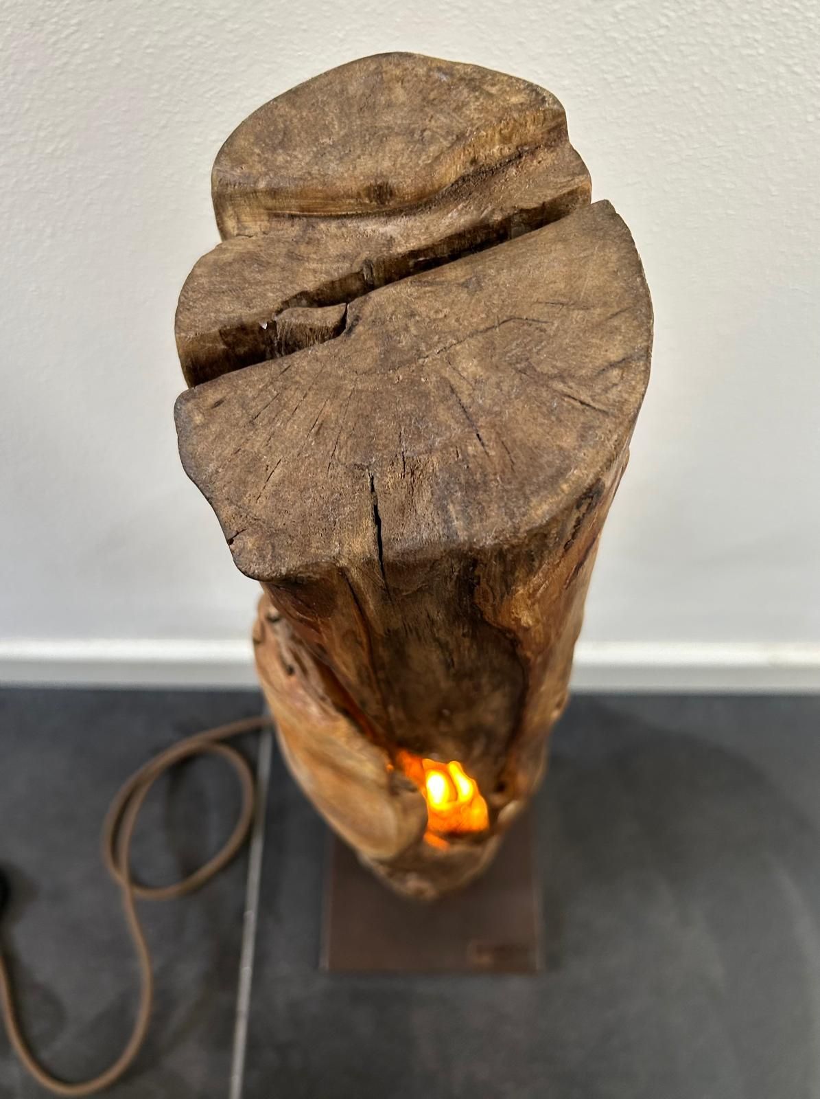 Lampada a sospensione con ceppo di legno e luce arancione, posizionata su un supporto con cavo e appoggiata a una parete spoglia.