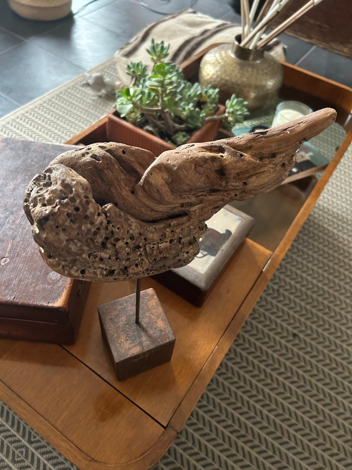 Scultura di uccello in legno su un piedistallo, esposta su un tavolo con piccole piante in vaso e decorazioni.