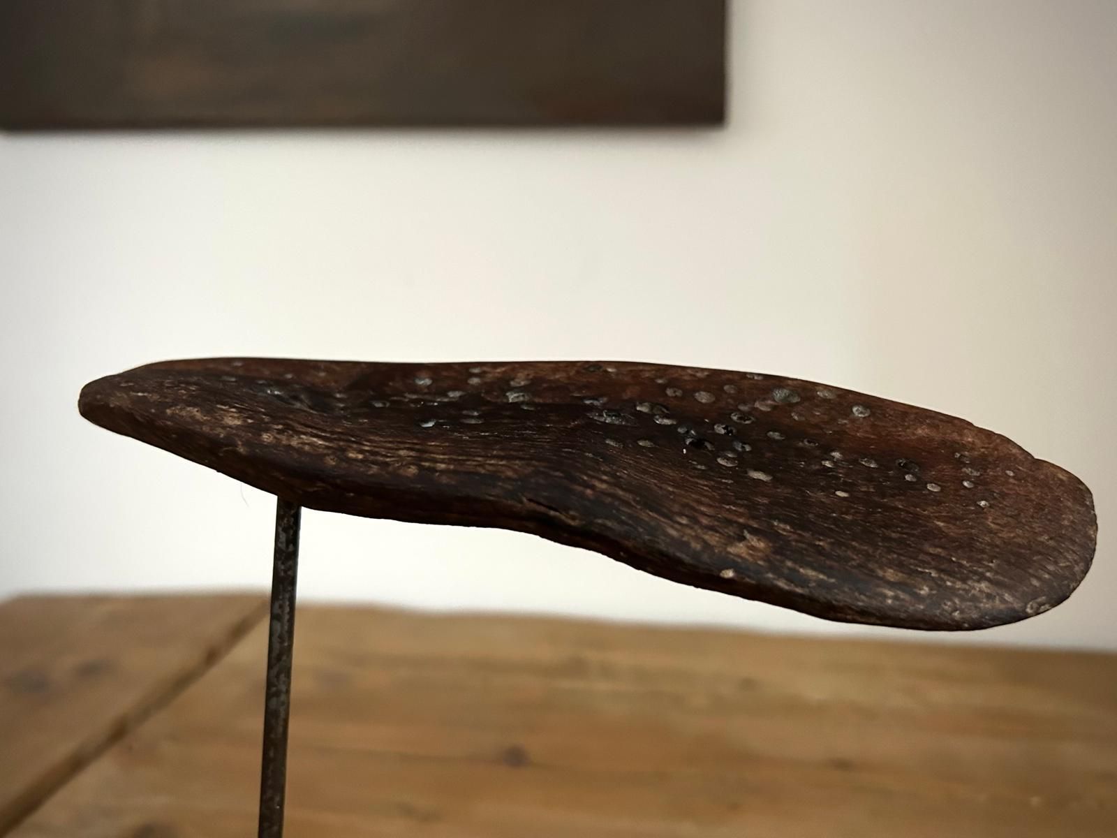 Oggetto intagliato in legno scuro su un sottile supporto metallico su sfondo chiaro.