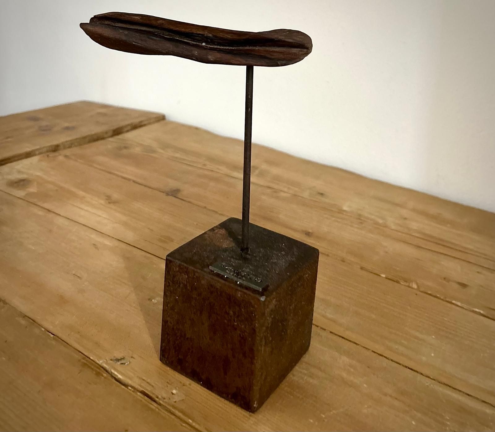 Piccola scultura astratta con una foglia di metallo scuro su un sottile stelo, posta su un blocco di legno appoggiato su un tavolo.