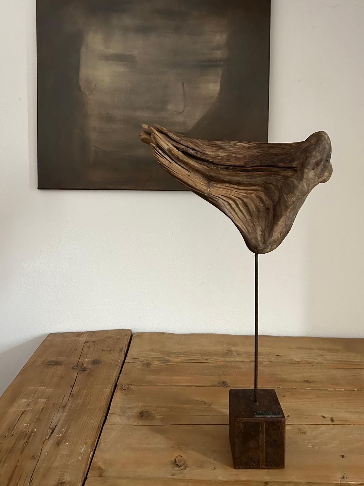 Scultura astratta in legno di recupero su un supporto metallico appoggiato su un tavolo di legno, con un dipinto dai colori tenui sulla parete retrostante.