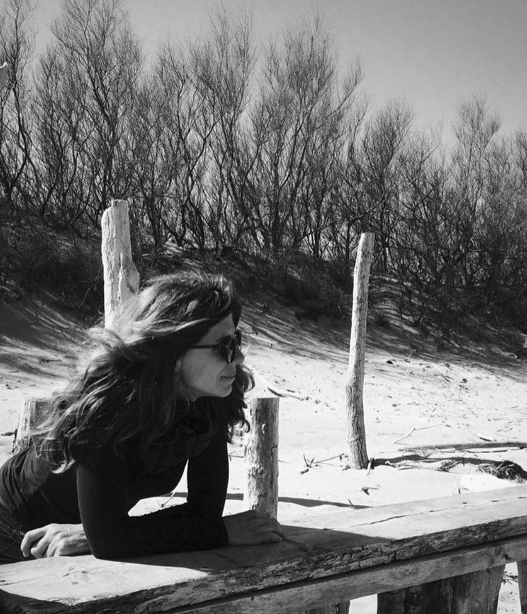 Persona sdraiata su una panchina di legno su una spiaggia sabbiosa, rivolta a sinistra, con alberi sullo sfondo.