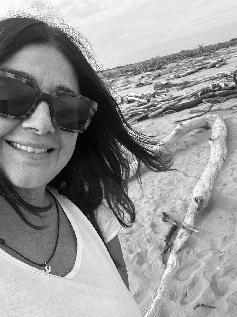 Una donna sorridente con gli occhiali da sole si scatta un selfie su una spiaggia sabbiosa con legni portati dalla corrente e onde sullo sfondo.