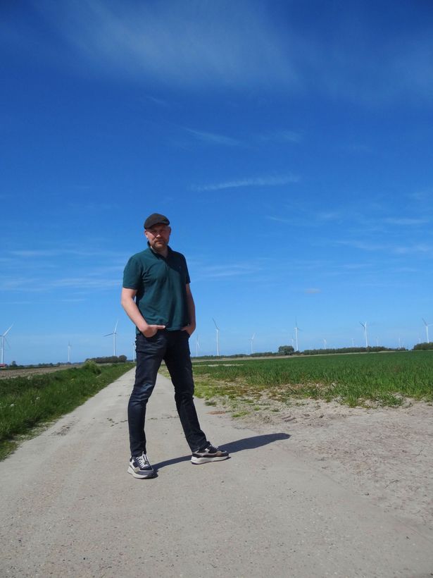 Een persoon staat op een zandpad in een grasveld onder een blauwe hemel, gekleed in een turquoise shirt en een donkere broek.