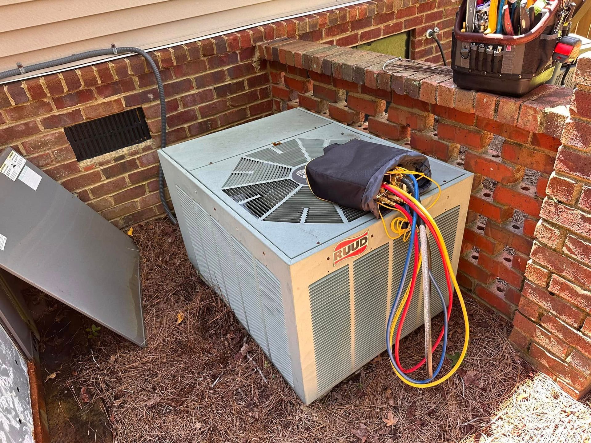 Outdoor HVAC unit beside brick wall with open panel, tools, and colored wires on top