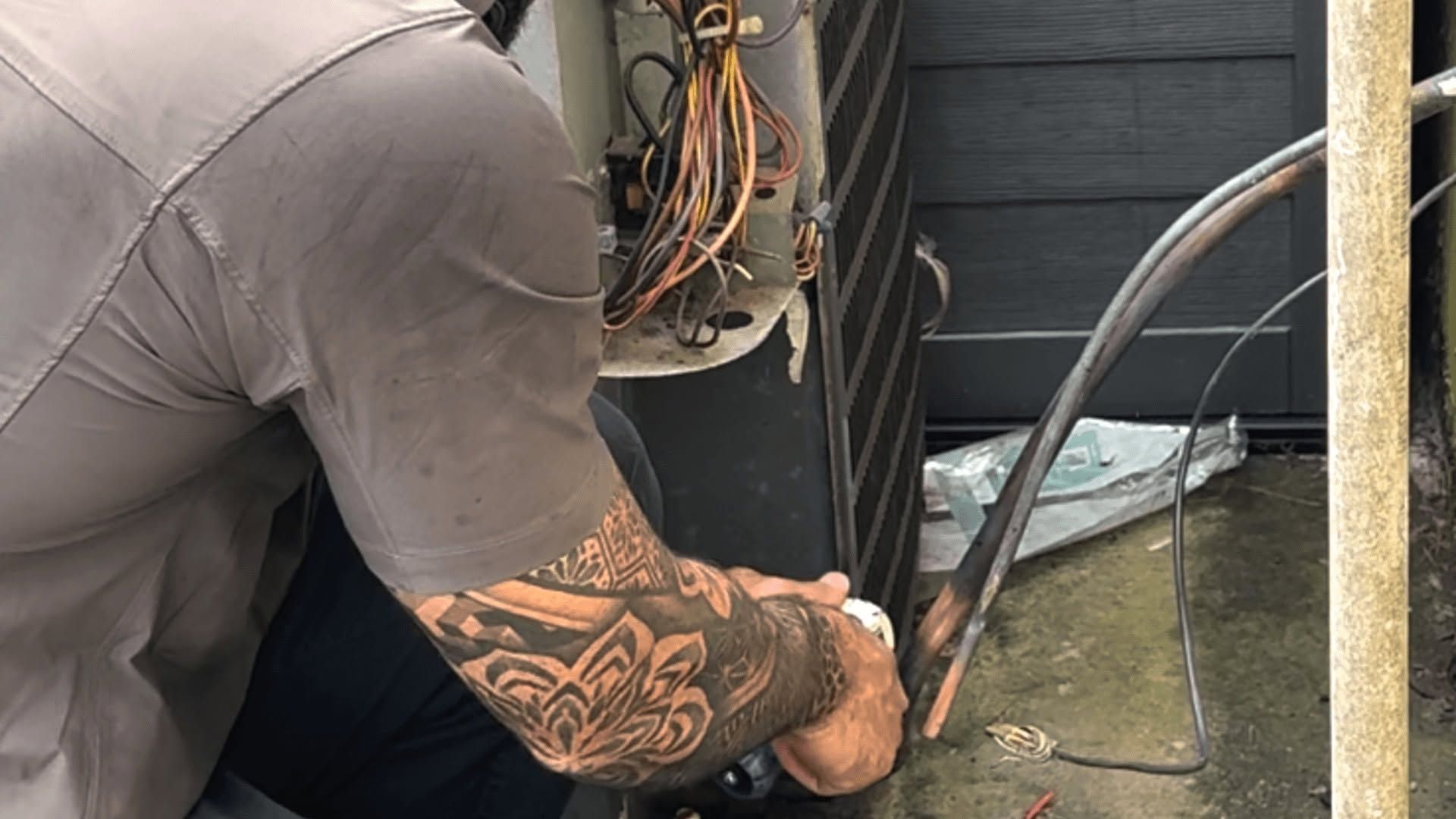 Technician kneeling beside outdoor HVAC unit, working on exposed wires with a tool