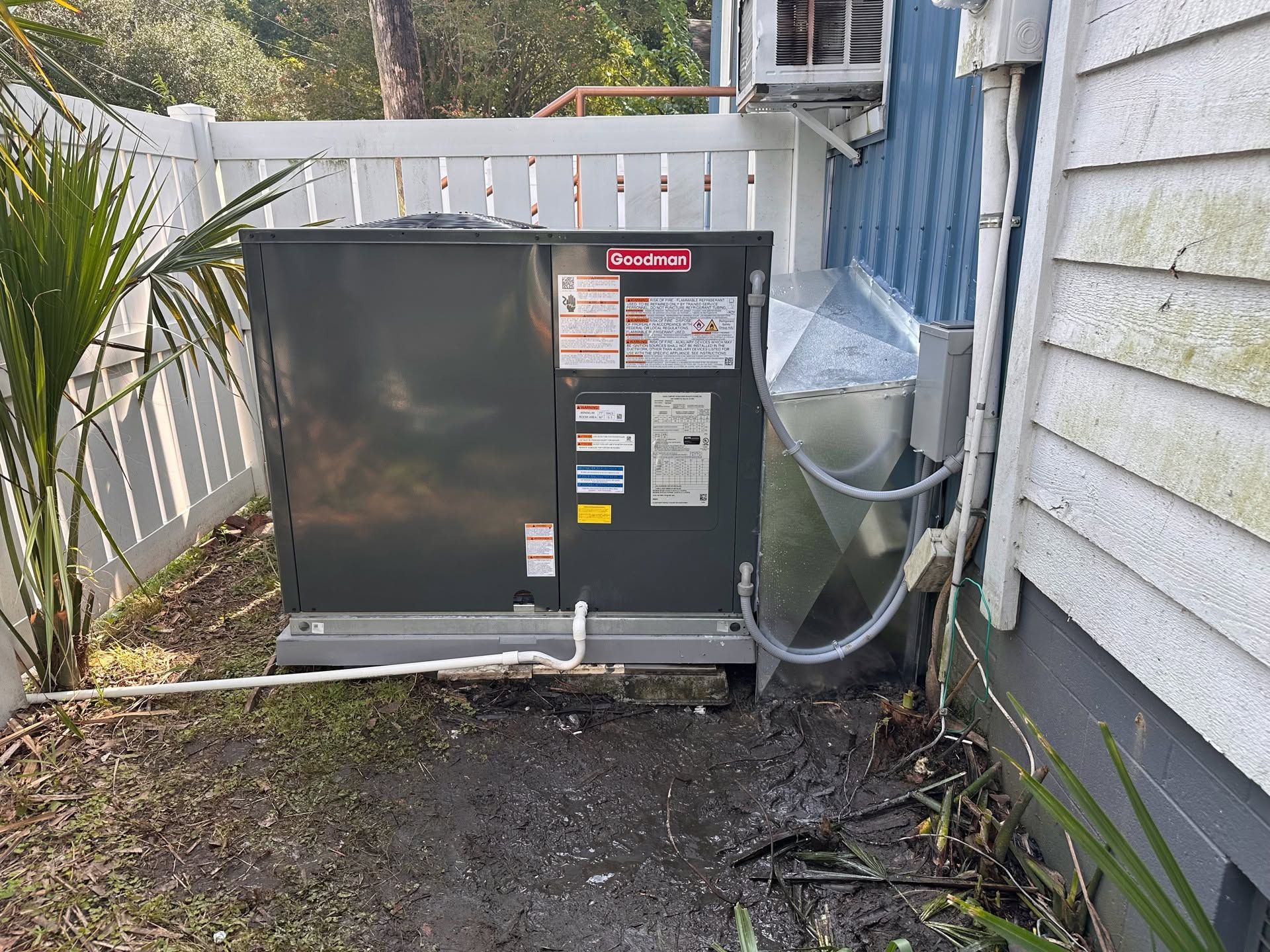 Outdoor HVAC unit beside a white house, with vents, pipes, and a clear cover near a gravel path.