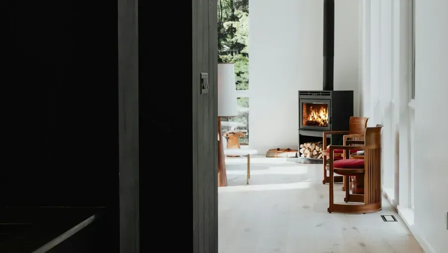 A minimalist hallway leading to a bright room with a small fireplace, chair, and wooden stool.