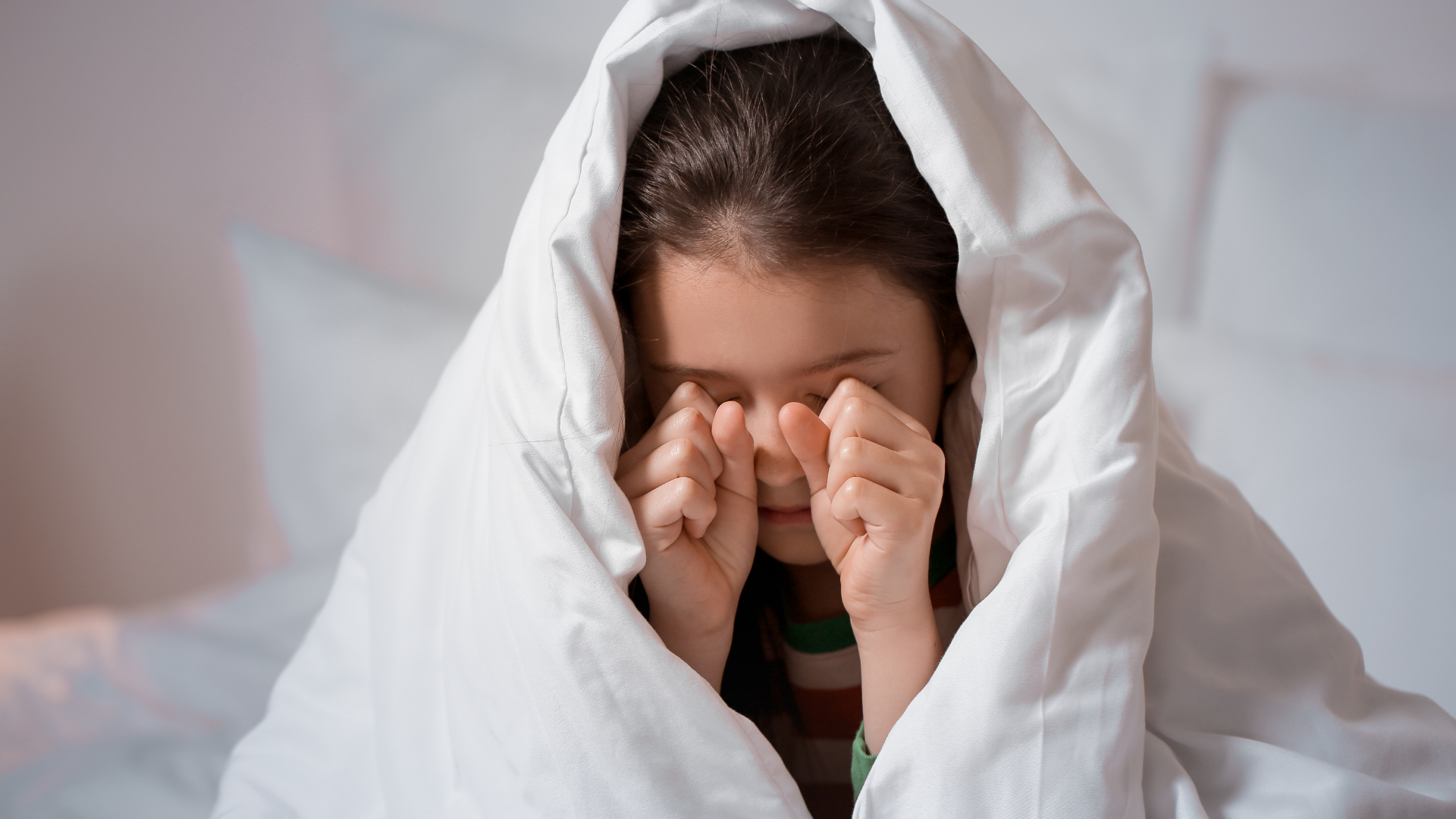 Girl in bed, rubbing eyes under a white blanket, appearing sleepy.