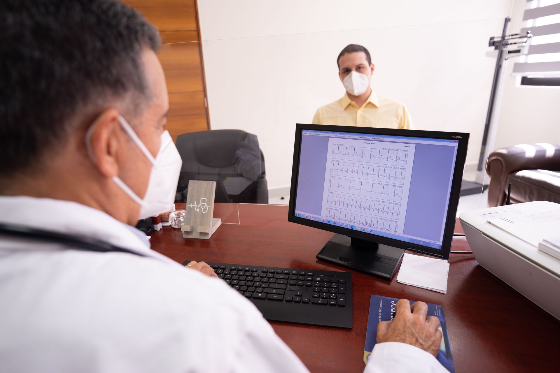 Doctor reviewing an x-ray on a monitor across the desk from a masked patient in a clinic office