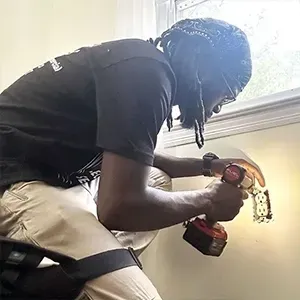 Person wearing glasses and a bandana installs a blue wall fixture with a drill in a room with a damaged ceiling.