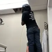 Person in black shirt and jeans working on a ceiling panel in a room with a ladder and exposed opening
