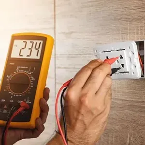 Hand holding multimeter probes to wall outlet, testing voltage reading 234 on digital display
