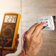 Hand holding multimeter probes to wall outlet, testing voltage reading 234 on digital display