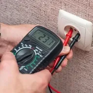Hand holding a multimeter testing a wall outlet with red and black probes, reading 230 volts