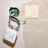 Exposed electrical wires and wall switch on a beige wall, with a loose control module hanging below.