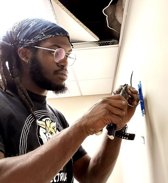 Person in glasses and bandana using a drill to install a blue wall fixture in a room with a ceiling opening