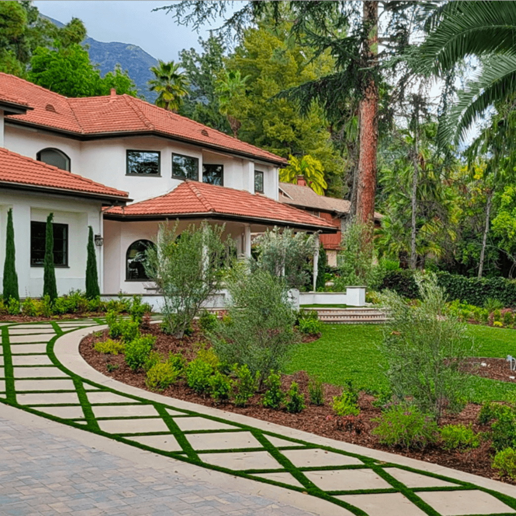 White house with red tile roof, curved driveway with green grass squares, lush landscaping.