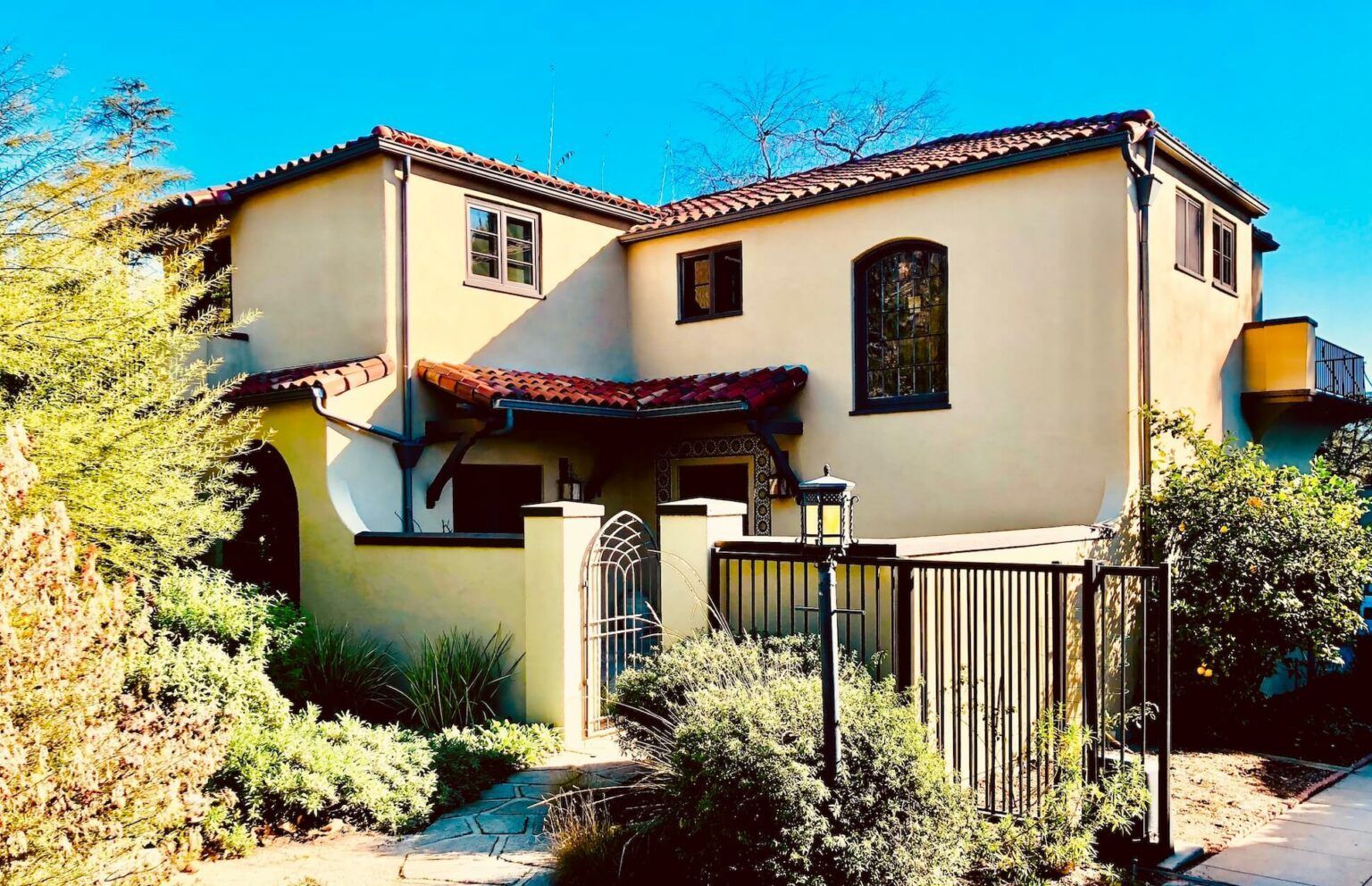 Beige stucco two-story house with a red tile roof and black iron gate. Lush greenery surrounds the house.