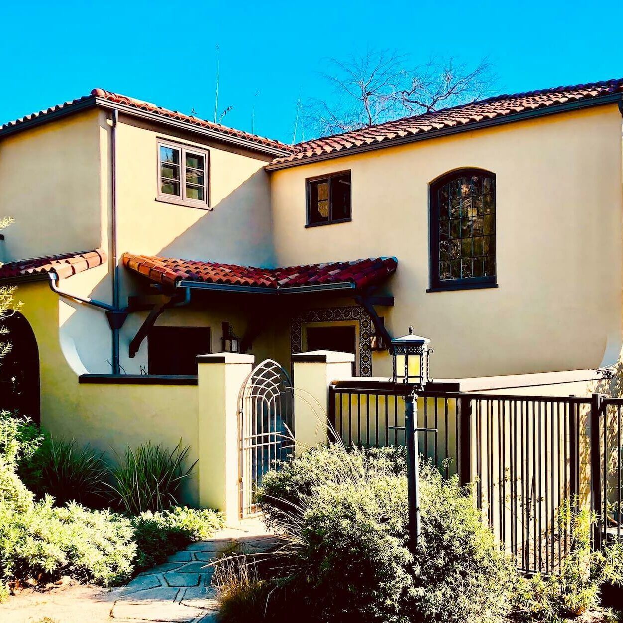 Beige stucco house with red tile roof, wrought iron gate, and lush landscaping under a sunny sky.
