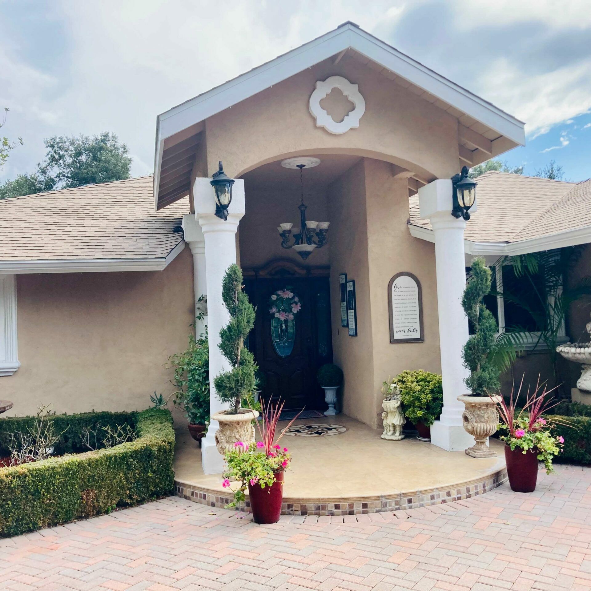 Tan house exterior with a brick driveway, ornate pillars, and vibrant flower arrangements.