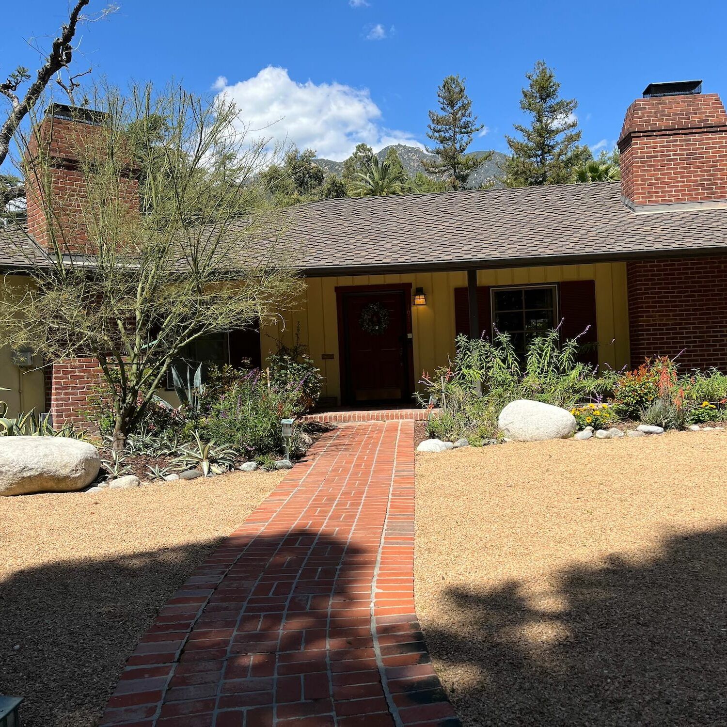 Red brick home with a path, yellow siding, and garden, mountains in background.