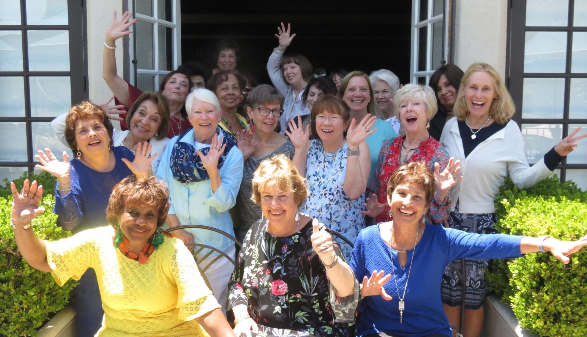 A group of smiling women wave from a doorway, sunny outdoors.