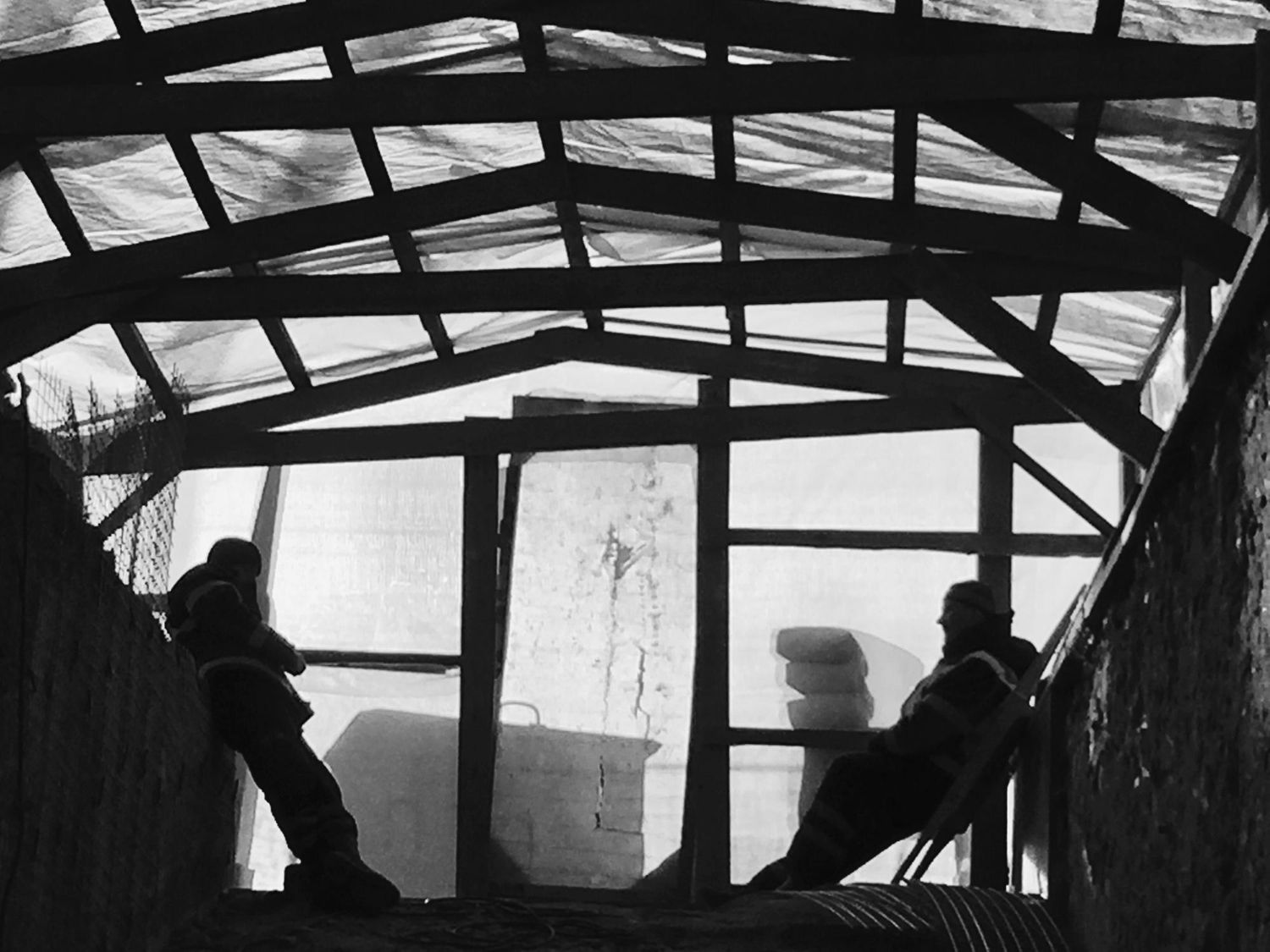 Zwei Personen auf einem Holzbalken unter einem Dachstuhl, als Silhouetten in einem baustellenartigen Innenraum erkennbar.