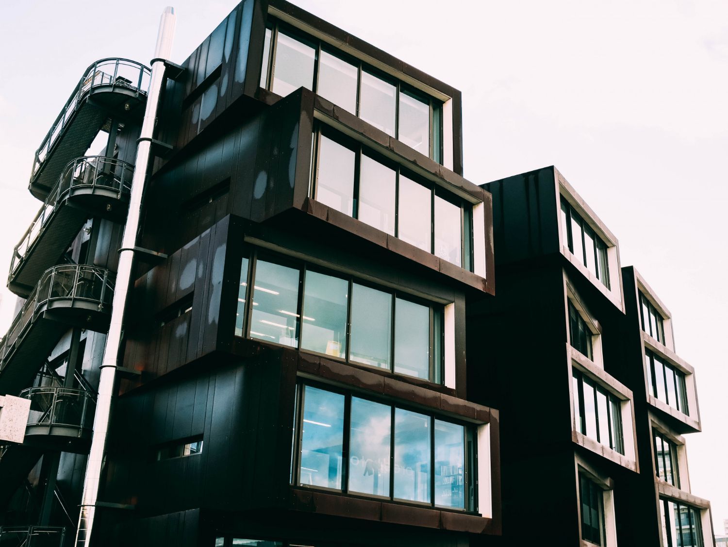 A low-angle view of a modern, multi-story building with dark, box-like modular sections and large windows.